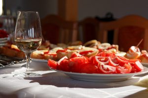 Tisch mit weisser Tischdecke, ein Weinglas mit Weisswein und ein weisser runder Teller voll mit geschnittener Tomaten