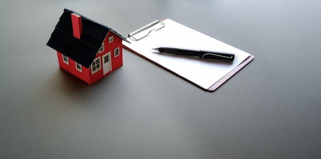 ein kleines rotes Spielzeughaus das neben einem Notizblock und einem schwarzem Kugelschreiber auf einem grauem Tisch steht